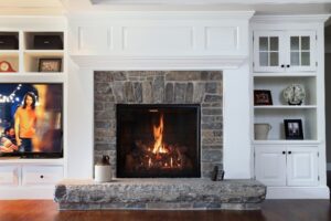 A Mendota gas fireplace surrounded by a stone hearth and white cupboards in a cozy room.
